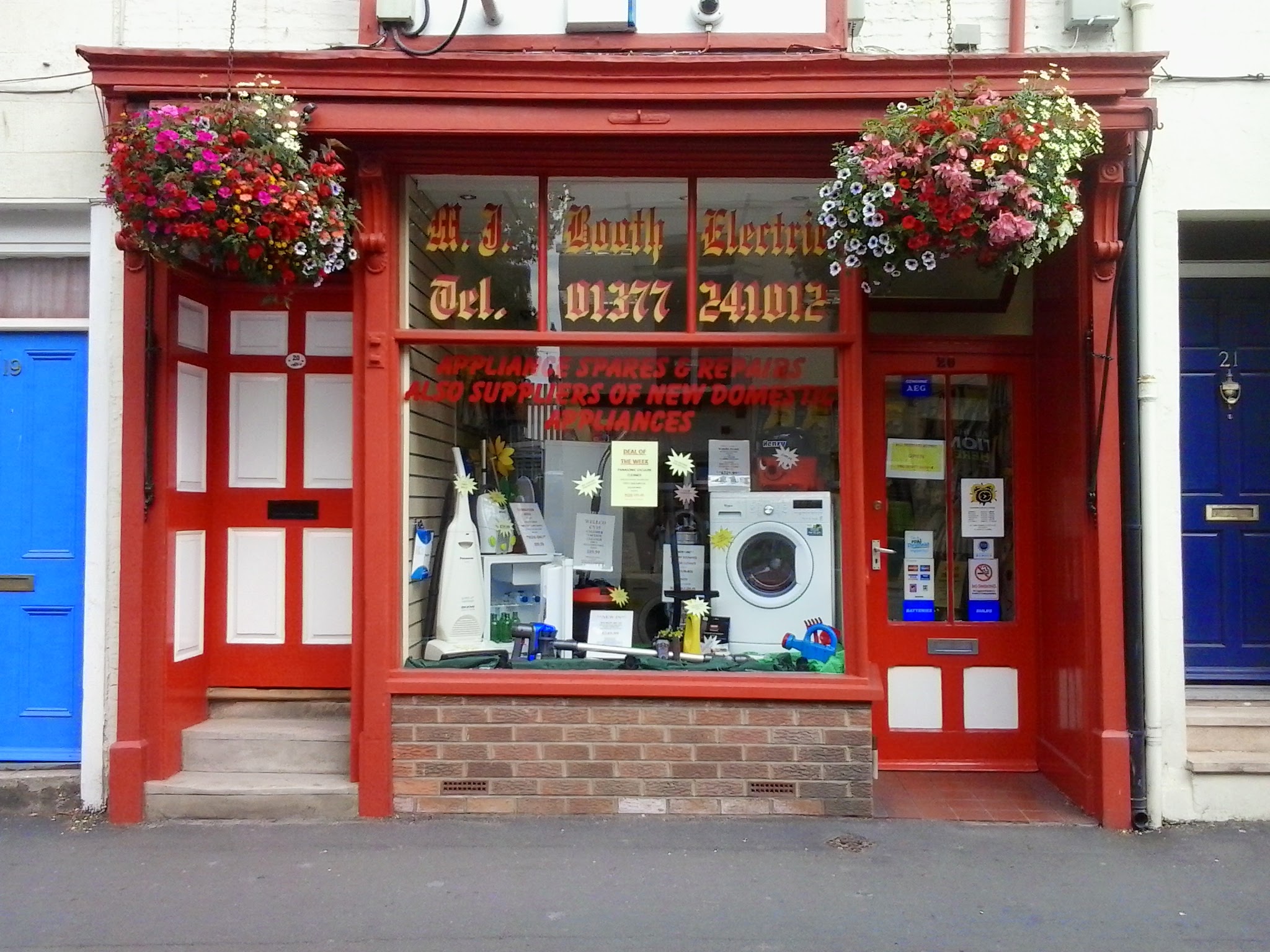 The M J Booth Electric shopfront painted red, with hanging baskets of flowers above the windows